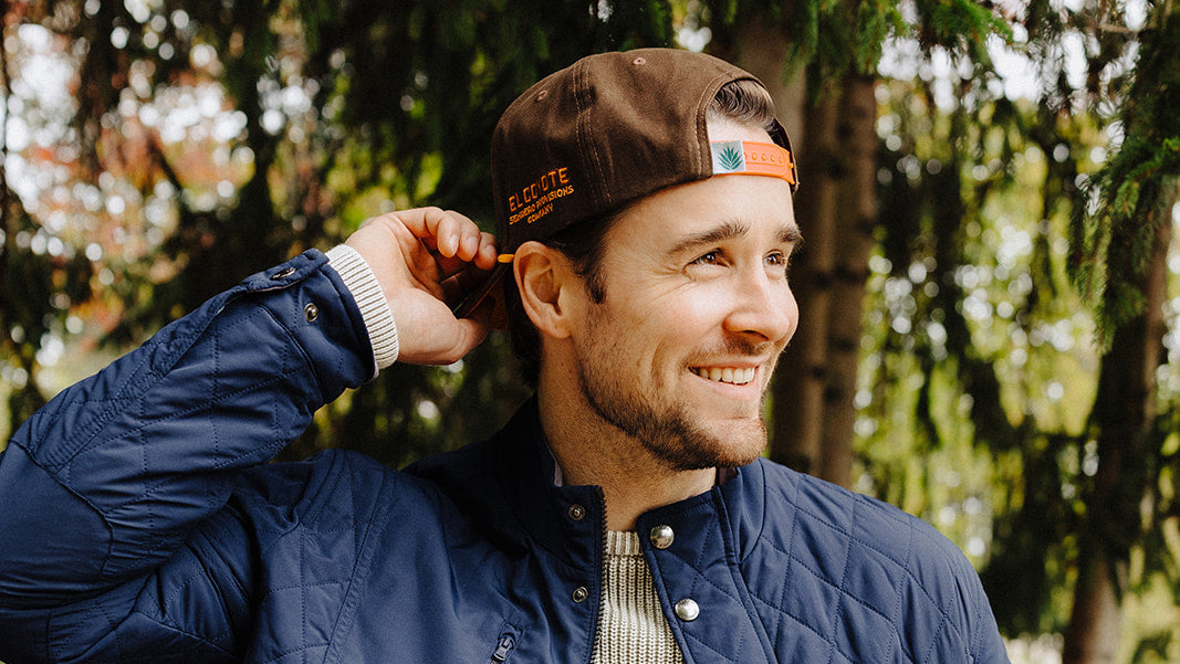 Man wearing a blue quilted jacket and brown cap outdoors with trees in the background